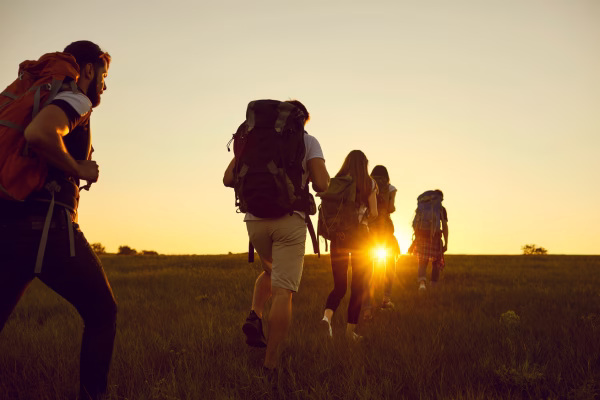 5 pessoas com mochilas grandes caminhando em um campo de grama ao por do sol